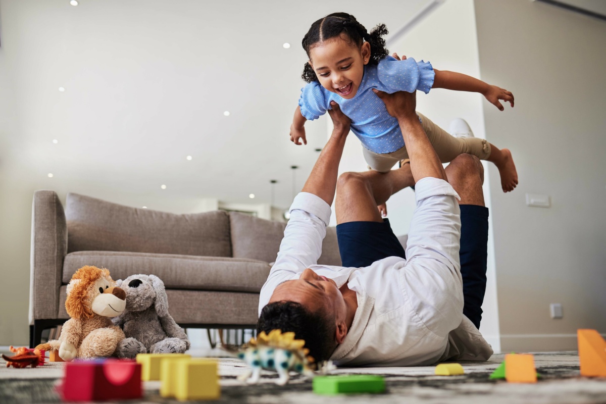 A father and young boy playing on the floor of the living room - the dad is lifting his son into the air with his hands and feet while on his back